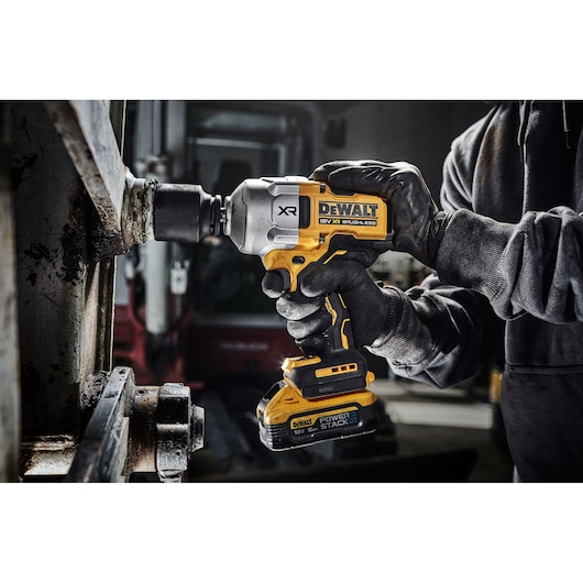 A person wearing gloves uses a DEWALT 18V XR Brushless impact driver with a POWERSTACK battery to work on a metal structure