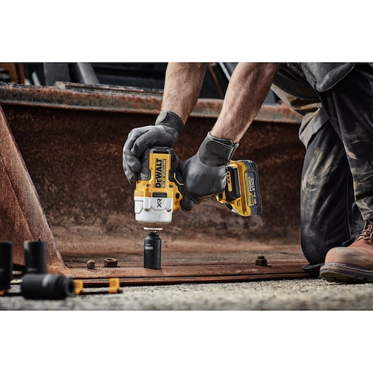A person wearing work gloves is using a DEWALT cordless impact wrench on a rusty metal plate, surrounded by bolts and