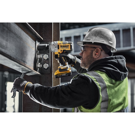 A construction worker wearing a safety helmet and reflective vest uses a DEWALT DCF961H2T cordless impact wrench to tighten