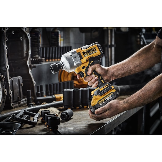 A person uses a DEWALT cordless impact wrench in a workshop with various sockets and tools on the bench.