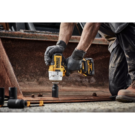 A person wearing work gloves uses a DEWALT cordless impact wrench to fasten or loosen a bolt on a metal plate at a construction site. The tool features yellow and black DEWALT branding.