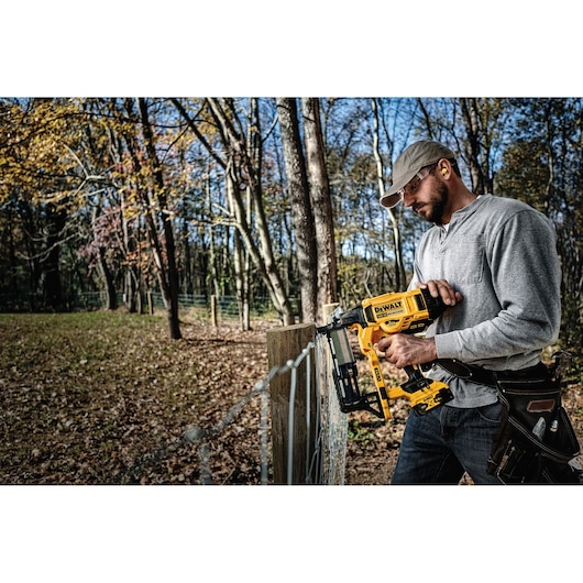 A person wearing a gray shirt and tool belt is using a DEWALT DCFS950P2 cordless fencing stapler to secure wire mesh to a