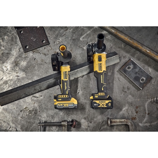 Two DEWALT cordless angle grinders on a metal workbench with tools and metal plates nearby.