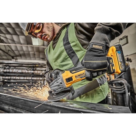 A person wearing safety gloves and a vest uses a DEWALT cordless angle grinder, model DCG405H2T, to cut metal, producing