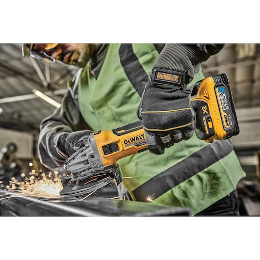 A person wearing protective gloves and a safety vest is using a DEWALT cordless angle grinder to cut metal, generating