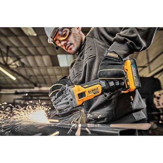 A person wearing gloves uses a DEWALT DCG405H2T cordless angle grinder to cut metal, with sparks flying, in an industrial