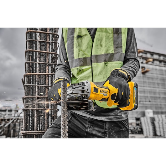 A person wearing a safety vest and gloves is using a yellow DEWALT cordless angle grinder to cut through a metal rebar at a