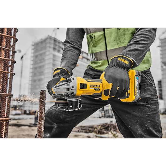 A worker wearing gloves and a safety vest uses a DeWalt cordless angle grinder to cut through a steel rod at a construction