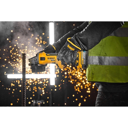 A person wearing safety gloves and a reflective vest uses a DEWALT cordless angle grinder to cut through a metal rod, with