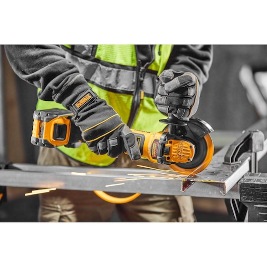 A person wearing protective gloves and a high visibility vest uses a DEWALT cordless angle grinder to cut a metal piece, with visible sparks flying.