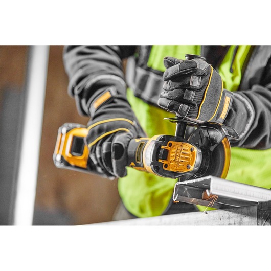 A person wearing protective gloves and a high-visibility vest is using a DEWALT cordless angle grinder to cut a piece of metal.