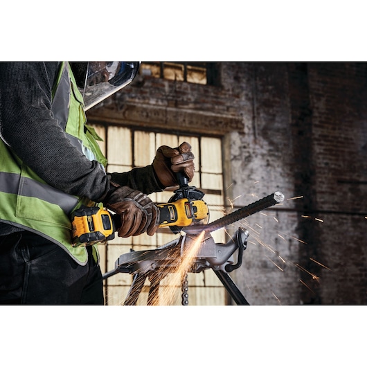 A person wearing protective gloves and a safety vest uses a yellow DEWALT cordless angle grinder to cut through a metal rebar, with sparks flying in an industrial workshop setting.