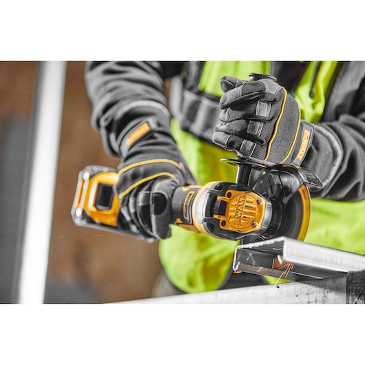 Close-up of a person wearing protective gloves and a high-visibility vest using a DEWALT cordless angle grinder to cut a piece of metal.