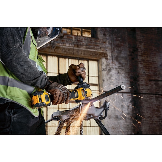 A person wearing safety gear and gloves uses a DEWALT cordless angle grinder to cut a metal rod, creating sparks in an