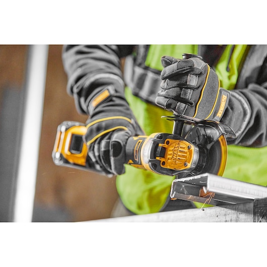A person wearing safety gloves and a high-visibility vest is using a DEWALT cordless angle grinder to cut a metal piece.