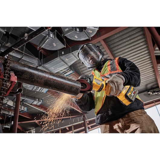 Person using DEWALT cordless grinder to cut pipe, wearing safety gear and generating sparks in industrial setting.