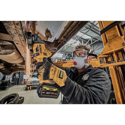 A person wearing protective gloves uses a DEWALT cordless angle grinder (model DCG418N) to cut or grind a metal component under a vehicle in a workshop. Sparks are visible from the grinding process.