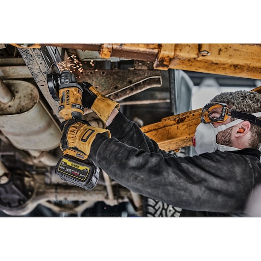 Person using a DEWALT cordless angle grinder (model DCG418N) to cut metal parts underneath a vehicle. The individual is wearing work gloves, safety goggles, and ear protection. Sparks are visible from the grinding process.