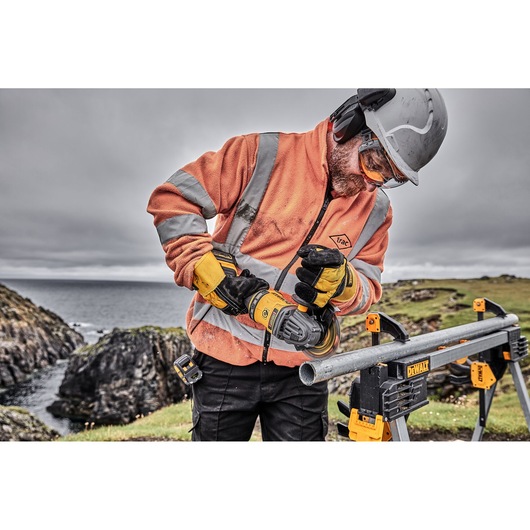 A worker wearing an orange safety jacket, gloves, helmet, and protective glasses is using a DEWALT power tool to cut a metal pipe outdoors. The pipe is supported by a DEWALT workbench, and the scene is set against a rocky coastline.