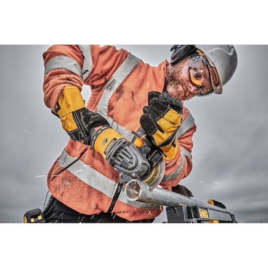 A person wearing safety gear and gloves uses a DEWALT cordless angle grinder to cut a metal pipe, with sparks visible during the process.