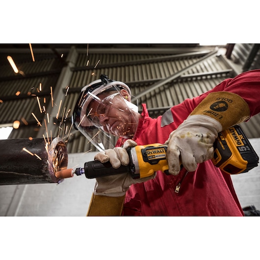 A person wearing protective gloves and safety gear uses a DEWALT DCG426N cordless grinder on a metal pipe, creating sparks in an industrial environment.