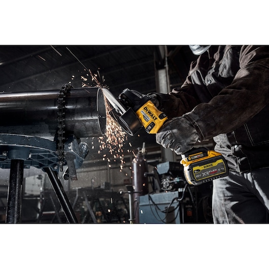 A worker uses a DEWALT cordless angle grinder to cut a metal pipe, with sparks flying in an industrial workshop setting.