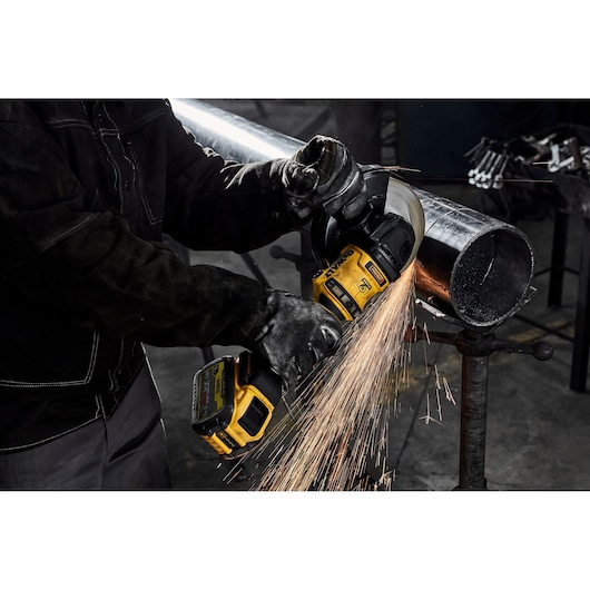 A person wearing protective gloves uses a yellow DEWALT DCG440N cordless grinder to cut a metal pipe, with bright sparks flying from the grinding wheel.