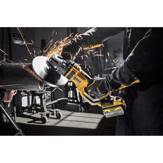 A person using a DEWALT cordless angle grinder to cut or grind a metal pipe, producing sparks. The tool is held with both