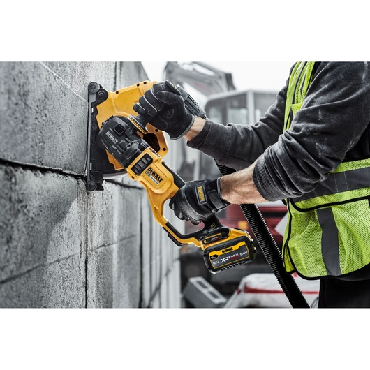 A person wearing gloves and a high-visibility vest uses a DEWALT cordless wall chaser tool to cut a straight line into a