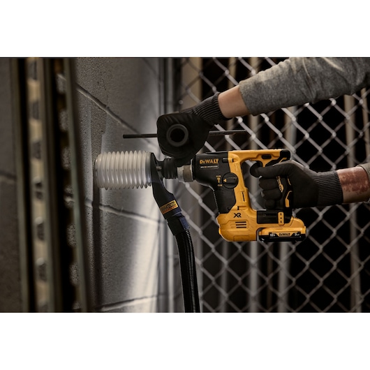 A person wearing gloves uses a DEWALT cordless rotary hammer drill (model DCH072L2) with a dust extraction attachment to drill into a concrete wall.
