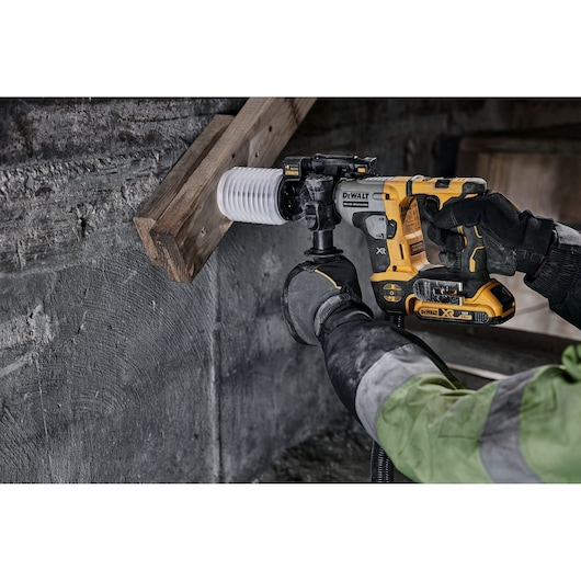 A person wearing gloves and a work jacket operates a yellow DEWALT cordless rotary hammer drill to make a hole in a wooden