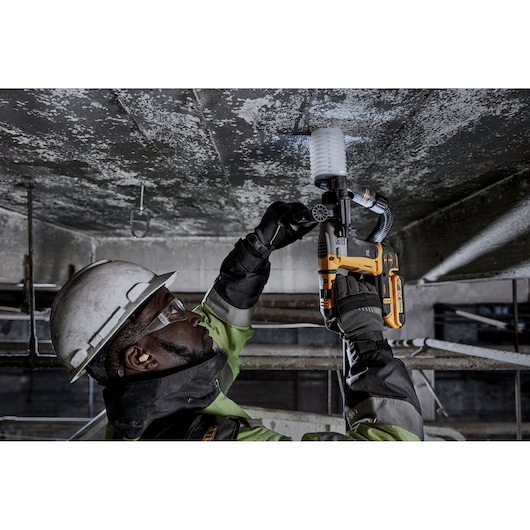 Person uses a DEWALT DCH172N rotary hammer drill on a ceiling, wearing safety gear and helmet at a construction site.