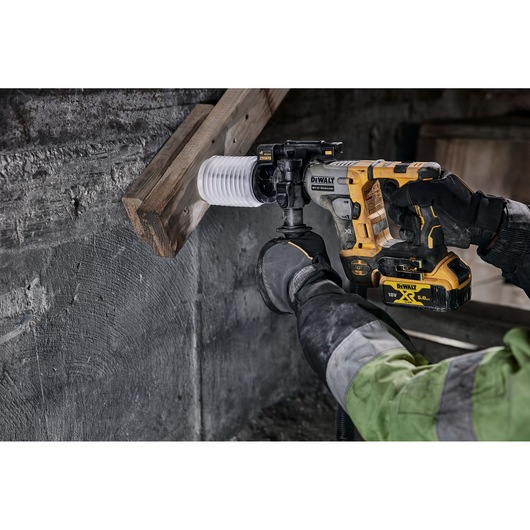 DEWALT cordless rotary hammer drilling into wood and concrete, operated by a gloved worker wearing a green jacket.
