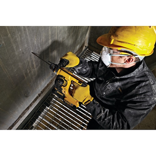 Worker uses a DEWALT DCH253N rotary hammer drill on a concrete wall, wearing safety gear and gloves.