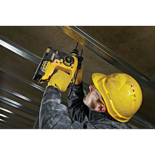 Person in safety gear uses a DEWALT DCH253N power tool to drill into a ceiling structure at a worksite.
