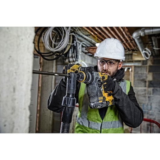 A person wearing a safety vest, gloves, and a hard hat is using a DEWALT rotary hammer drill (model DCH274P2T) to work on a