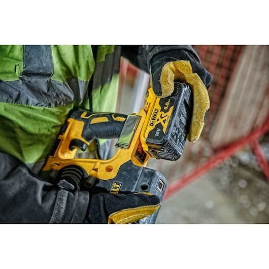 A close-up of a person wearing work gloves and a safety jacket, holding a DEWALT cordless power tool with an 18V XR lithium