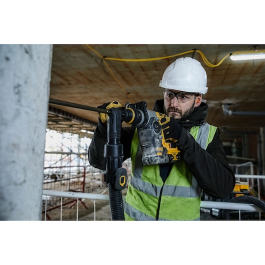 A person wearing a safety helmet and high-visibility vest operates a DEWALT DCH274P2 rotary hammer drill at a construction site.