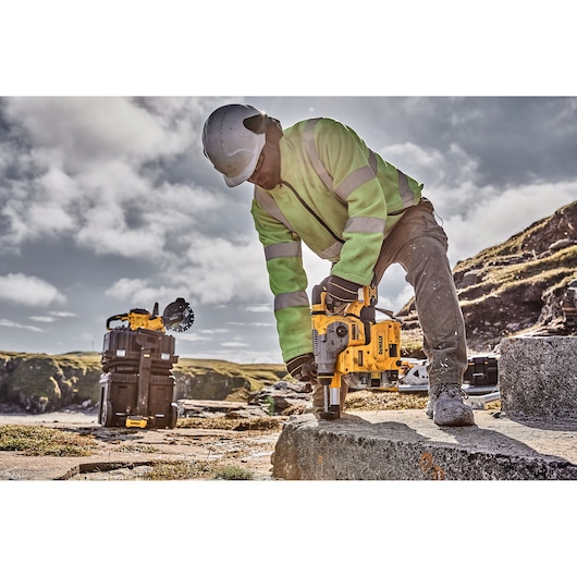 A person wearing a safety helmet and high-visibility jacket is using a DEWALT DCH283P2 rotary hammer drill on a concrete surface outdoors. Another DEWALT tool and storage boxes are seen in the background.