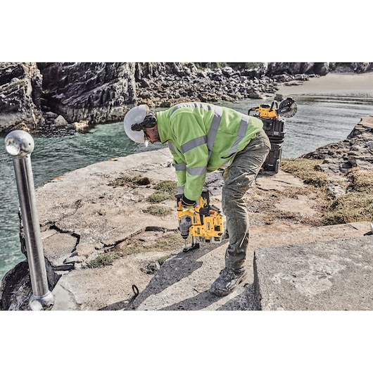 A construction worker wearing protective gear and a high-visibility jacket using a yellow DEWALT rotary hammer drill on a cracked concrete surface near water, with DEWALT tools and equipment in the background.