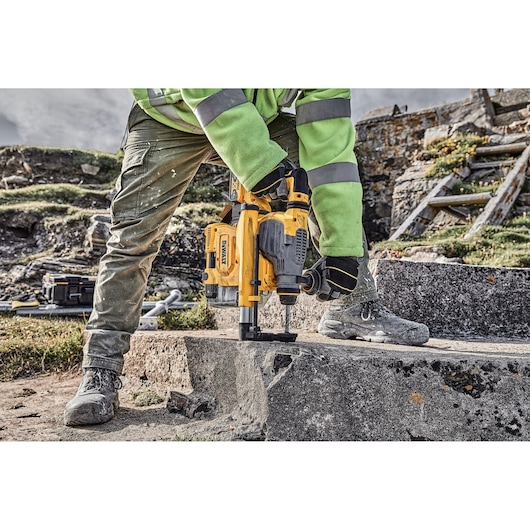 A person wearing work clothing is using a yellow DEWALT rotary hammer drill to drill into a concrete surface outdoors. Construction tools and equipment are visible in the background.