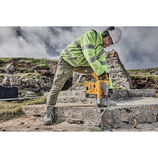 A person wearing safety gear, including a helmet, ear protection, a high-visibility jacket, and work boots, operates a DEWALT cordless rotary hammer drill (model DCH283P2) on a concrete slab outdoors at a construction site.