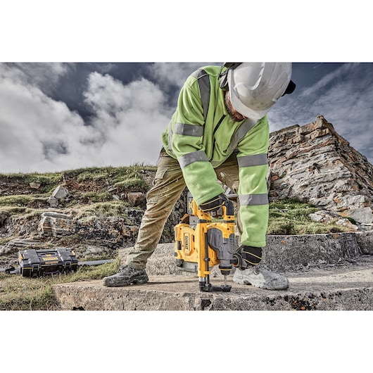A person wearing a high-visibility jacket and a hard hat is using a yellow DEWALT rotary hammer drill on a concrete surface outdoors, with a rocky landscape and a DEWALT toolbox visible in the background.