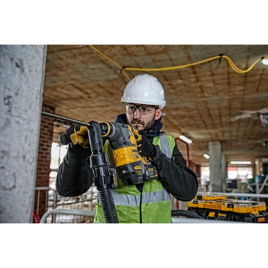 A construction worker wearing a hard hat, safety vest, and gloves is operating a DEWALT rotary hammer drill (DCH323T2) on a