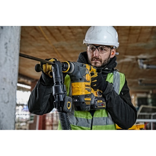 A worker wearing a safety helmet, gloves, and a high-visibility vest is using a DEWALT DCH323T2 cordless rotary hammer drill