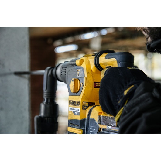 A DEWALT DCH323T2 cordless rotary hammer drill in use, held by a person wearing black gloves, drilling into a concrete wall.