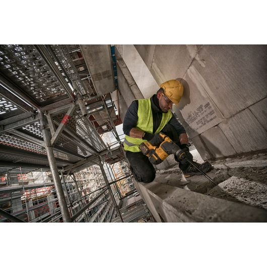 A construction worker wearing a yellow hard hat and high-visibility vest uses a DEWALT cordless rotary hammer drill to work