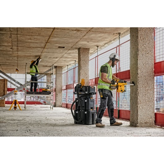 Two construction workers are operating DEWALT power tools at a building site. One worker is drilling into a concrete pillar, wearing a hard hat, safety vest, and protective gear, while the other worker is standing on scaffolding measuring the ceiling. DEWALT toolboxes and equipment are visible nearby.