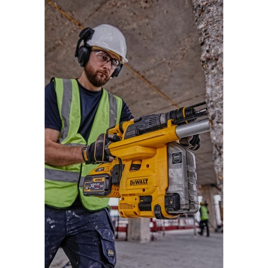 A person wearing safety gear and a high-visibility vest is holding a DEWALT DCH334X2 cordless rotary hammer drill with a battery, working at a construction site.