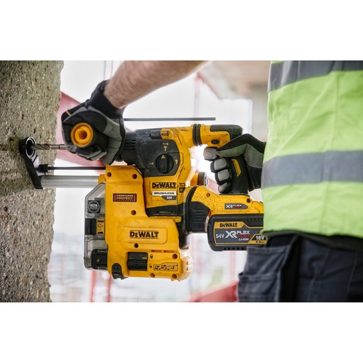 A person wearing gloves and a high-visibility vest is using a yellow DEWALT DCH334X2 cordless rotary hammer drill with a brushless motor to drill into a concrete wall.